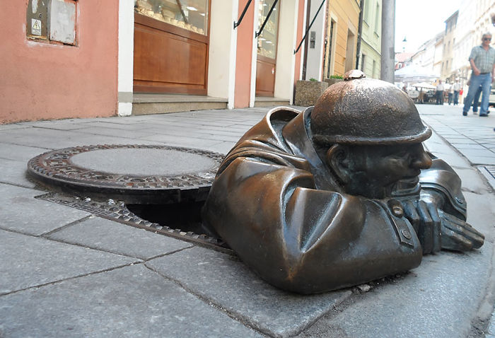 Bronze sculpture of a man crawling out of a manhole on a city sidewalk, showcasing amazing urban art.