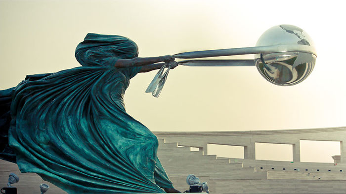 Amazing sculpture of a cloaked figure holding a reflective globe with long rods in an open, modern outdoor setting.