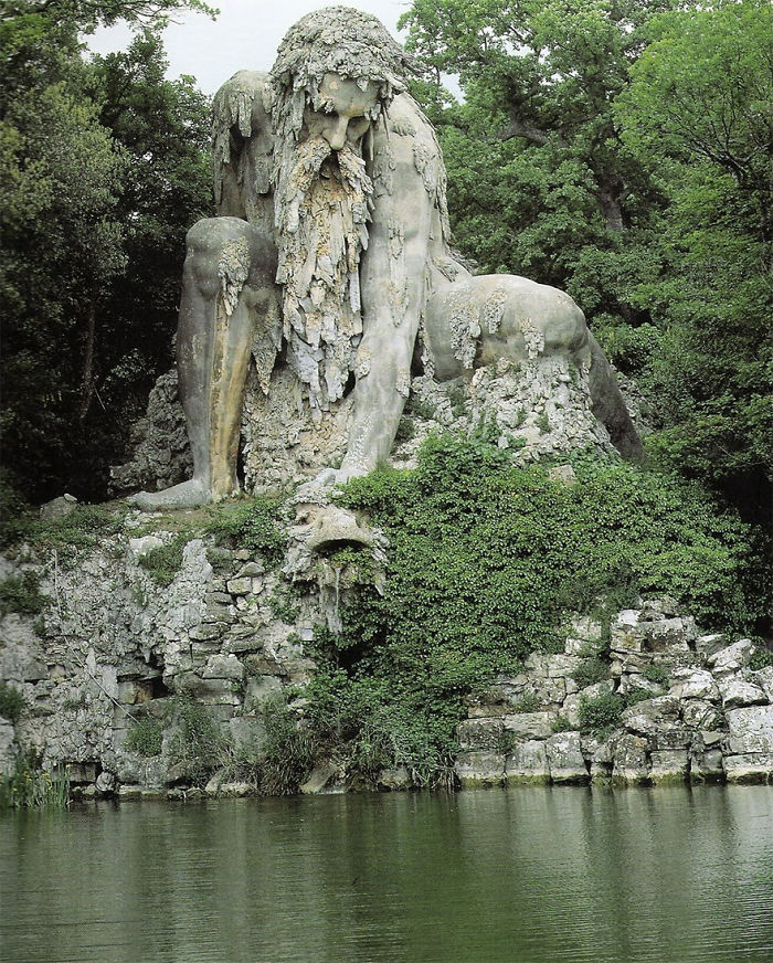 Large stone sculpture of a seated bearded figure covered in moss and vines beside a tranquil pond, nature surrounding it.
