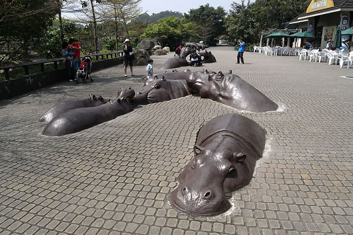 Large hippo sculptures appear to be emerging from a cobblestone walkway, showcasing amazing sculpture art outdoors.