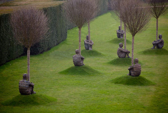 Sculptures of seated human figures integrated with trees on a grassy lawn in an outdoor art installation.