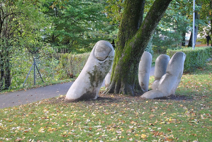 Large stone hand sculpture partially buried around a tree in a green park, one of the most amazing sculptures in the world.
