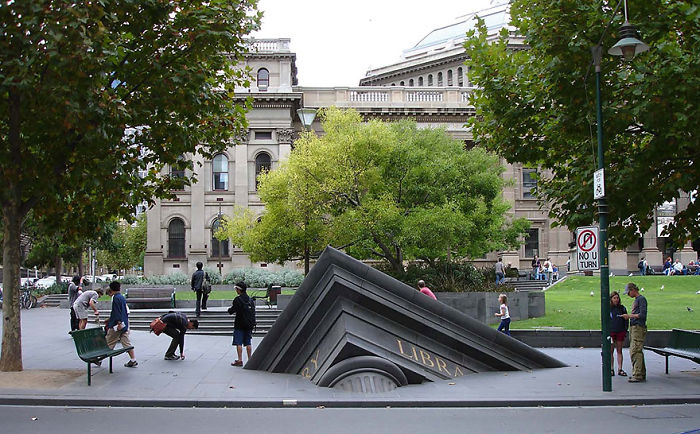 Sculpture of a sinking classical building with columns in an outdoor urban park surrounded by trees and people.