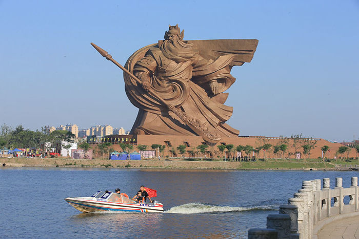 Large amazing sculpture of a warrior holding a spear near a river with a boat passing by under clear blue sky.