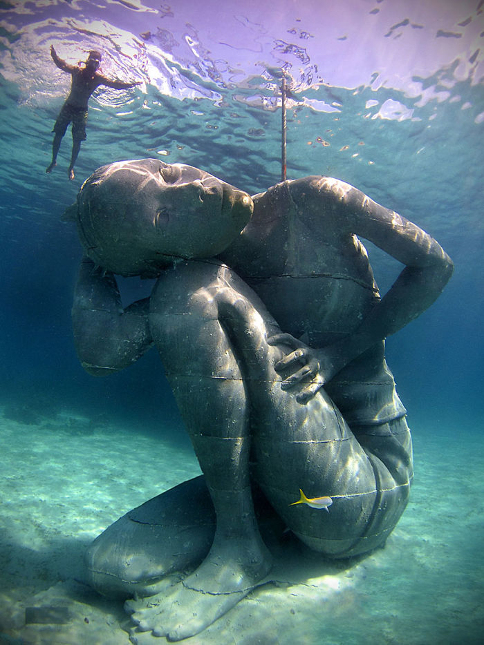 Underwater sculpture of a seated person with one hand behind the head, surrounded by clear water and a small fish nearby.