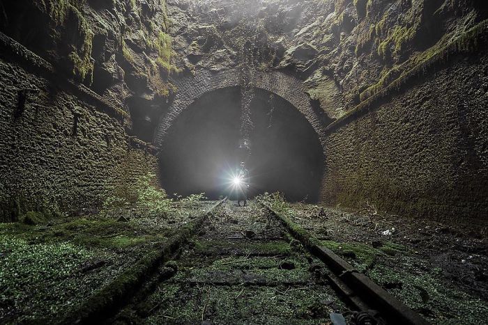 Person with flashlight exploring an abandoned railway tunnel covered in moss and vegetation in Britain’s abandoned places.