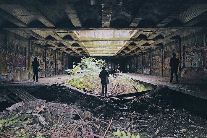 Three people explore an abandoned place in Britain covered with graffiti and overgrown plants in a decaying urban setting.