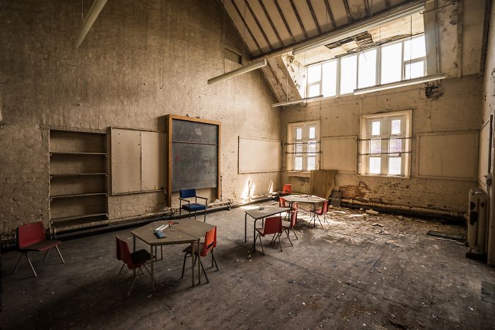 Abandoned Britain classroom with old desks, scattered chairs, and sunlight streaming through large dusty windows in an abandoned place.