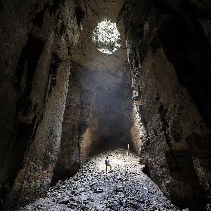 Person exploring a vast abandoned place in Britain, light streaming through an opening in the rocky ceiling.