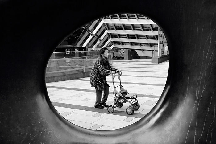 Black and white Japan street photography showing an elderly woman with a walker framed by a circular sculpture in an urban setting