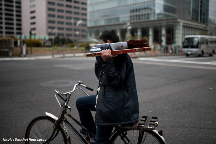 Man carrying trays on shoulder while riding bicycle on an urban street in Japan street photography shot