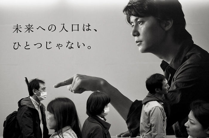 Black and white Japan street photography showing pedestrians with masks passing by a large mural of a man pointing.