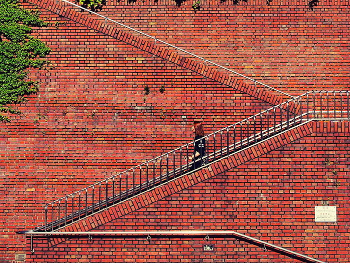 Woman walking up an outdoor brick staircase against a red brick wall in a Japan street photography shot.