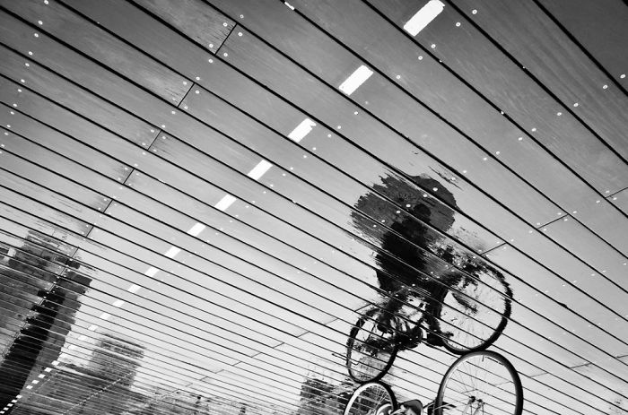 Black and white Japan street photography showing a distorted reflection of a cyclist on wet wooden planks.