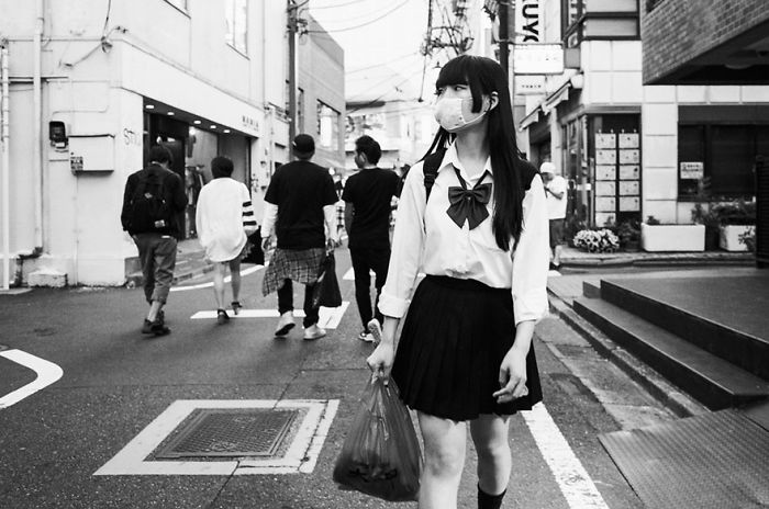 Black and white Japan street photography featuring a masked schoolgirl walking through a quiet urban street scene.