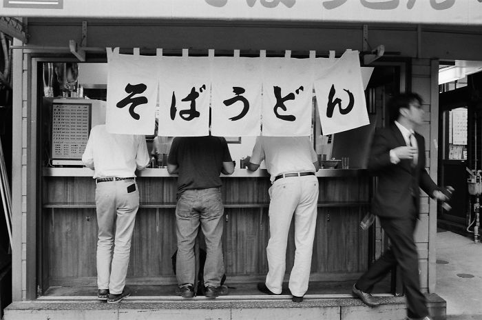 Black and white Japan street photography showing people eating at a small noodle shop in an urban setting.