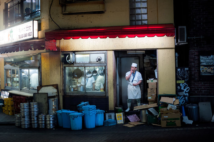 Japanese street photography capturing a chef outside a dimly lit restaurant with kitchen utensils and blue containers nearby.