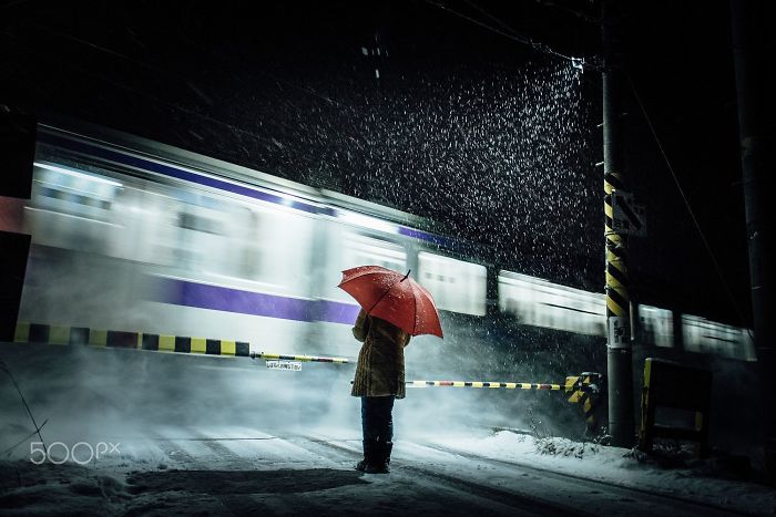 Person with red umbrella standing in rain by railroad crossing as a train passes, showcasing Japan street photography.