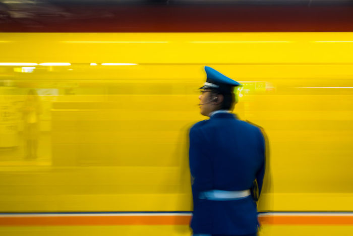 Japanese street photography showing a transit officer standing against a blurred yellow train in an urban setting.