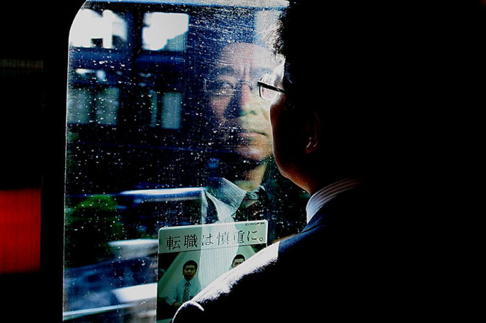 Man in glasses reflected on rainy window capturing Japan street photography with a dark, moody urban background.