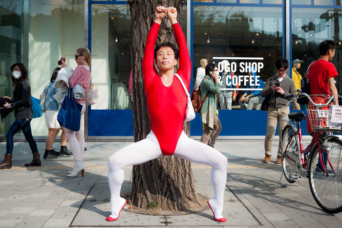 Man in red and white outfit stretching against a tree on a busy street in Japan street photography scene.
