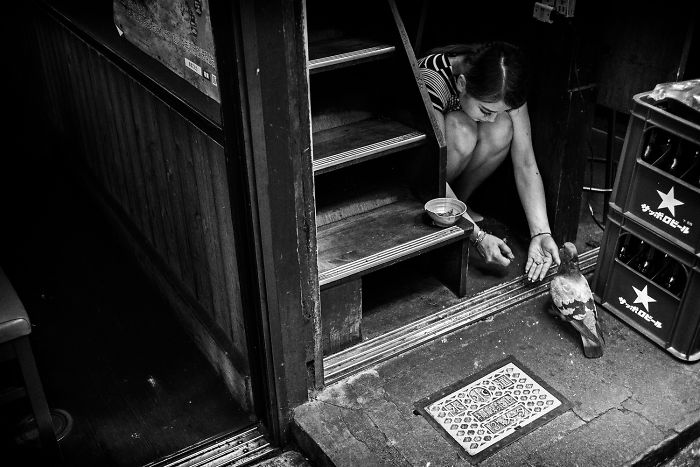 Black and white Japan street photography shot of a woman feeding a pigeon near a wooden doorway.
