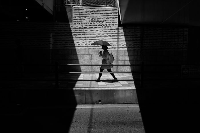 Black and white Japan street photography showing a person walking with an umbrella in a geometric light and shadow setting.