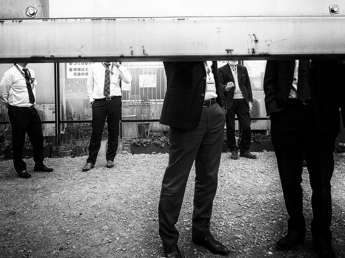 Men in suits standing behind a large metal beam captured in Japan street photography with a stark black and white style.