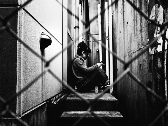 Black and white Japan street photography of a person sitting on stairs behind a fence in a narrow urban alleyway.