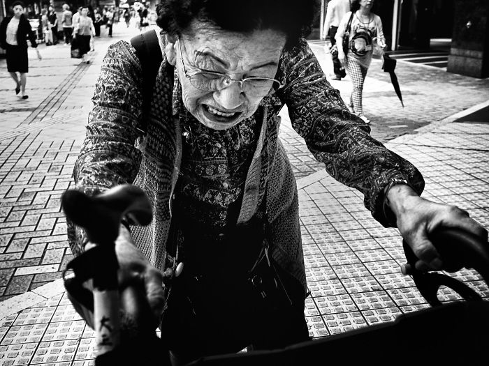 Black and white Japan street photography shot of an elderly woman pushing a bicycle on a busy city sidewalk.