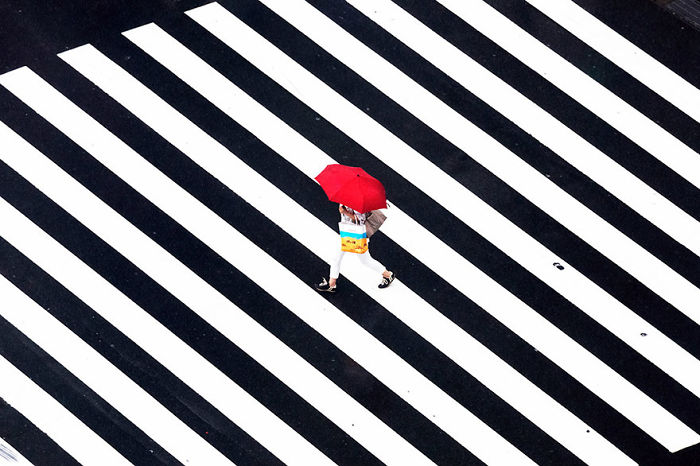 Person walking across a large black and white striped crosswalk in Japan street photography with a bright red umbrella.
