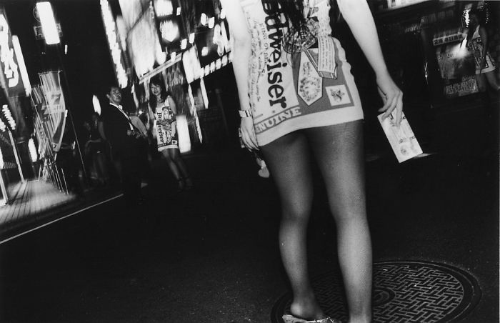 Black and white Japan street photography showing a woman in patterned dress and fishnet stockings on a city sidewalk at night.