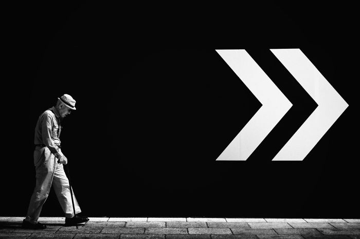 Black and white Japan street photography of an elderly man walking past a wall with large white arrows.