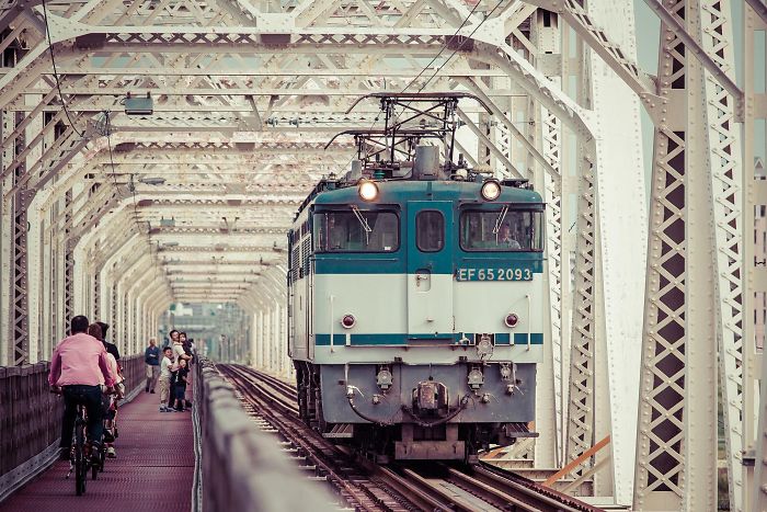 Train crossing an iron bridge with cyclists and pedestrians alongside in Japan street photography shot