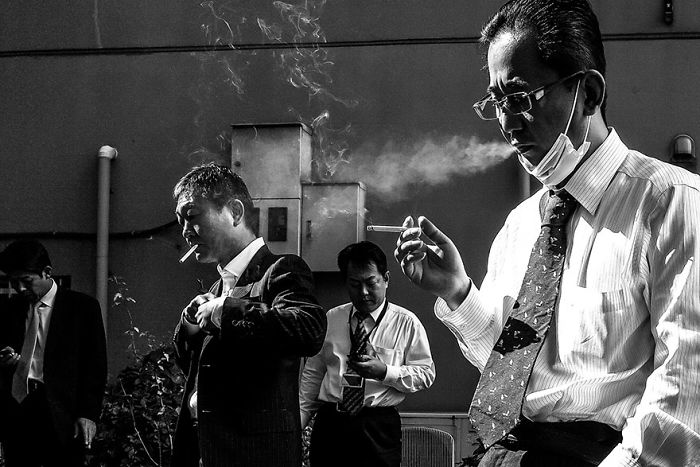 Black and white Japan street photography showing men in suits smoking and checking phones in a rare candid moment.