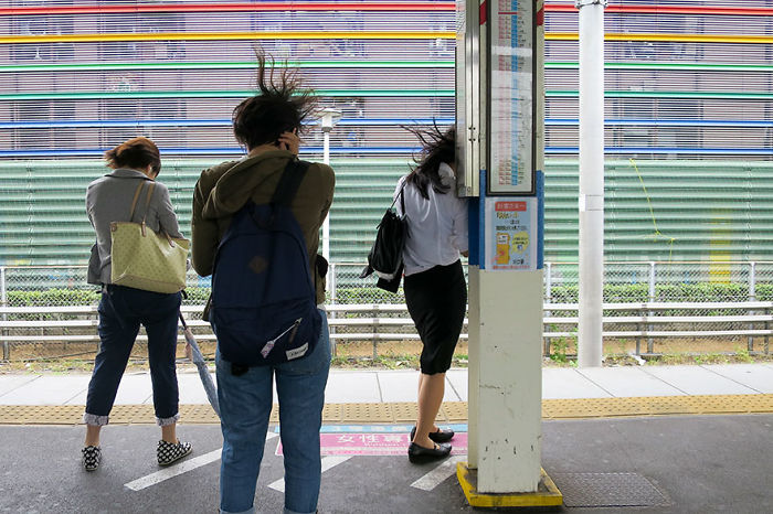 Three people with windswept hair waiting at a Japan street photography scene near a colorful striped wall and train platform.