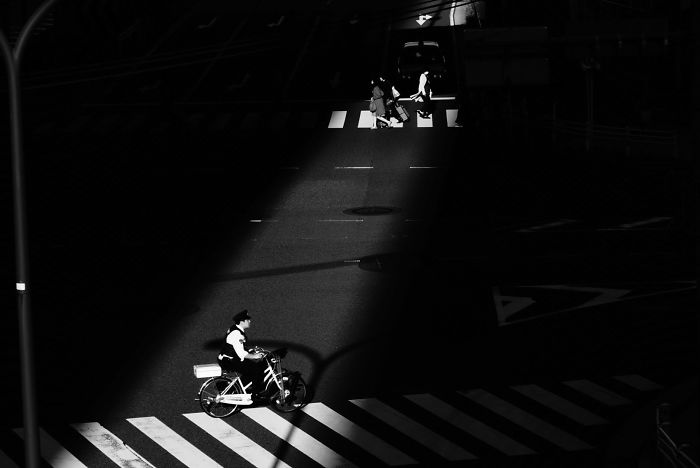 Black and white Japan street photography showing a cyclist and pedestrians crossing in a rare light and shadow scene.
