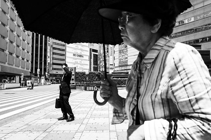 Black and white Japan street photography showing an elderly woman with umbrella and a man standing at a busy crosswalk.