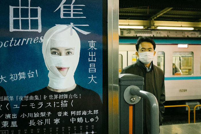 Japan street photography shot of a man wearing a mask standing near a subway train and a poster with a person wrapped in bandages.
