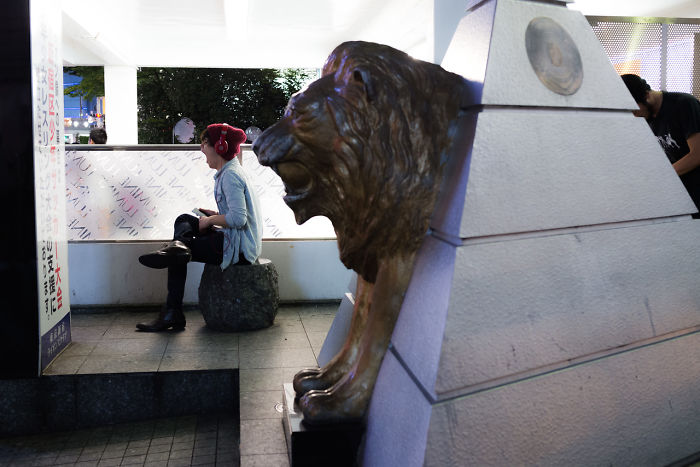 Person wearing red headphones sitting near a bronze lion statue in a city setting, showcasing Japan street photography.