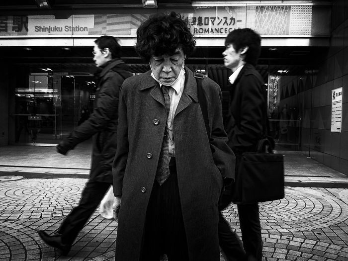 Black and white Japan street photography showing a contemplative man in a coat with blurred pedestrians at Shinjuku Station.