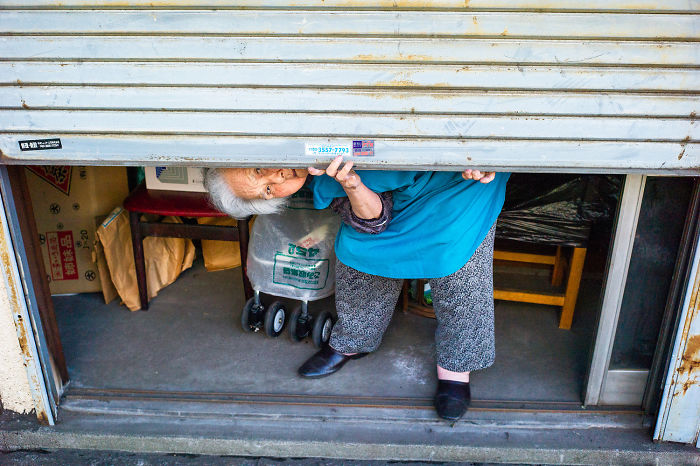 Elderly woman peeking from beneath a metal shutter in a narrow urban alley, Japan street photography scene.