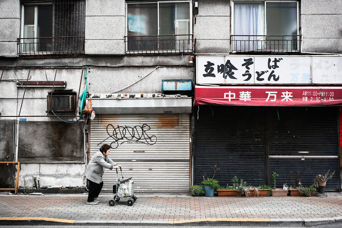 Elderly person walking with a cart in front of closed shops in a Japan street photography shot capturing everyday life.