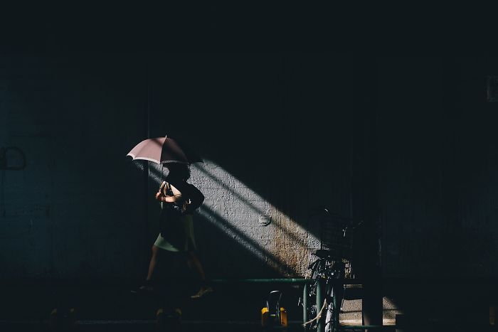 Person with an umbrella walking through a dark alley illuminated by a single light beam in Japan street photography.