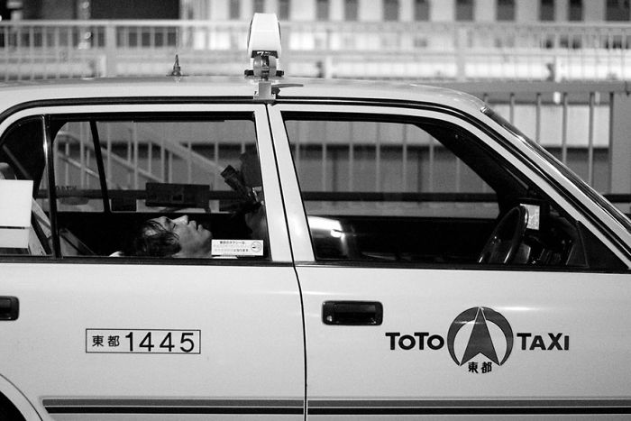 Black and white Japan street photography showing a taxi with a driver resting inside on a city street at night.