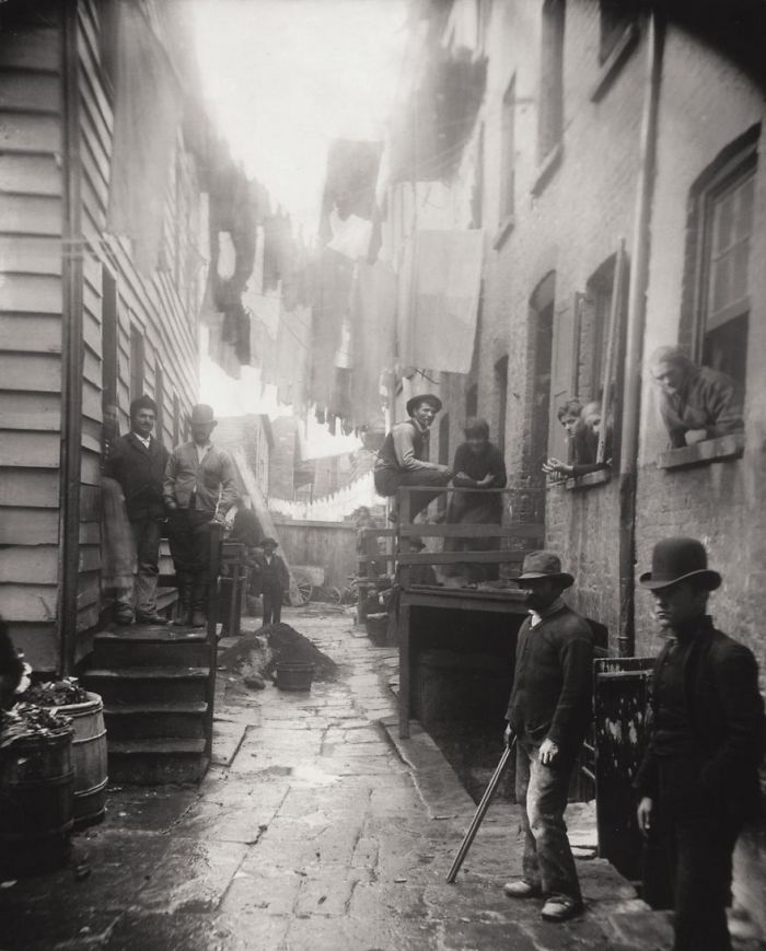 Historic photo of a crowded alleyway with people standing and sitting, laundry hanging overhead, capturing life in the past.