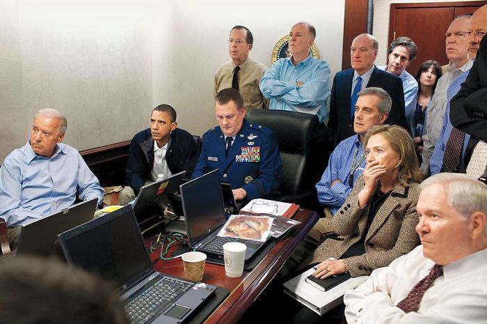 Group of officials in a meeting room focused on a historic event, with laptops and papers scattered on the table.