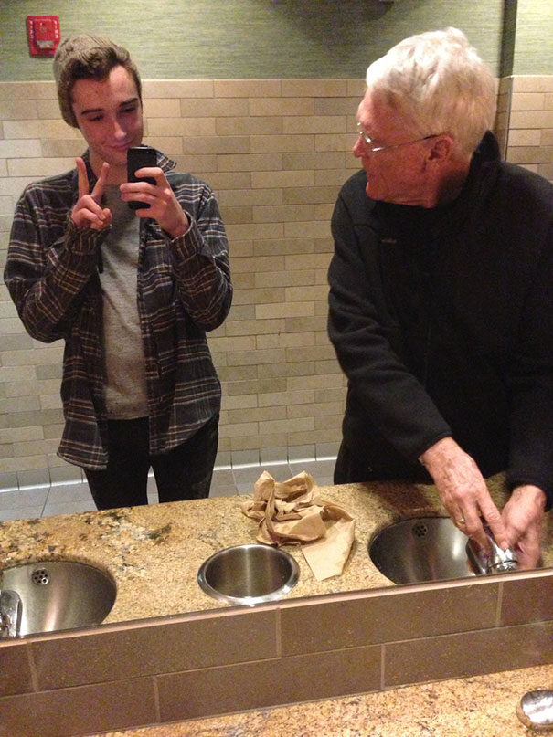 Boy making mirror selfie while old man washing hands 