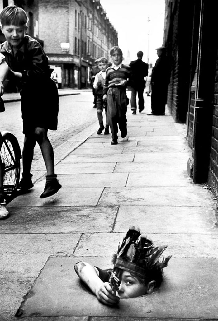 A Young Boy Wearing An Indian Headdress Hides In A Coal Hole And Takes Aim With A Toy Pistol, London, 1954