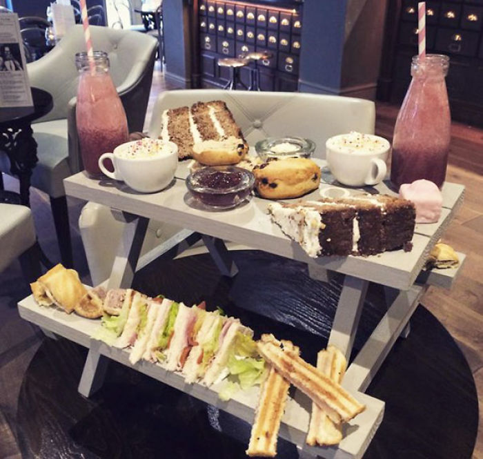 Elaborate restaurant food presentation with sandwiches, cakes, and drinks on a mini picnic table display.